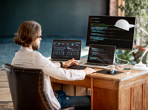 Developer working at a wooden desk with laptop screens showing software dashboards and code.